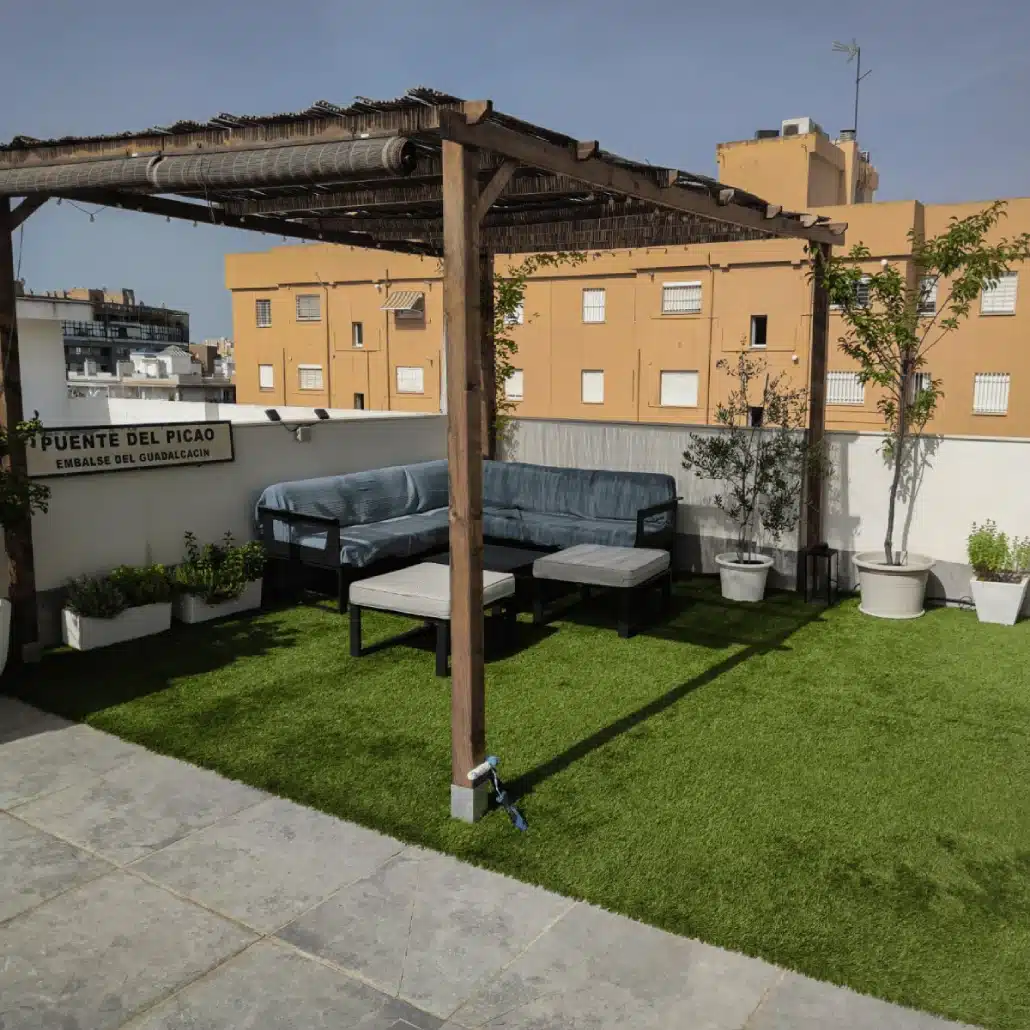Una terraza en la azotea con Césped artificial ADRIÁTICO 30mm, una pérgola de madera, un conjunto de sofás de exterior grises, plantas en macetas y edificios residenciales amarillos de fondo bajo un cielo despejado.