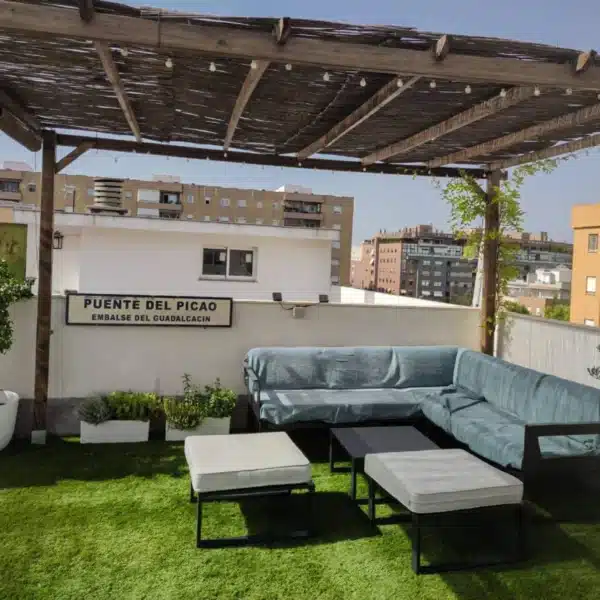 Una terraza en la azotea cuenta con Césped artificial ADRIÁTICO 30mm, asientos al aire libre con sofá y otomanas, una pérgola, plantas en macetas y un cartel PUENTE DEL PICAO EMBALSE DEL GUADALCACÍN. Al fondo se ven edificios de la ciudad.